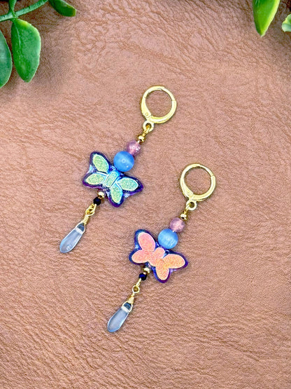 Handmade earrings featuring butterfly charms in blue and pink, with gold-tone hoops and clear crystal drops, displayed on a textured brown leather background with green leaves.