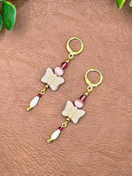 Handmade earrings with butterfly charms, pink faceted beads, and white teardrop beads, displayed on a textured brown leather background with green leaves.