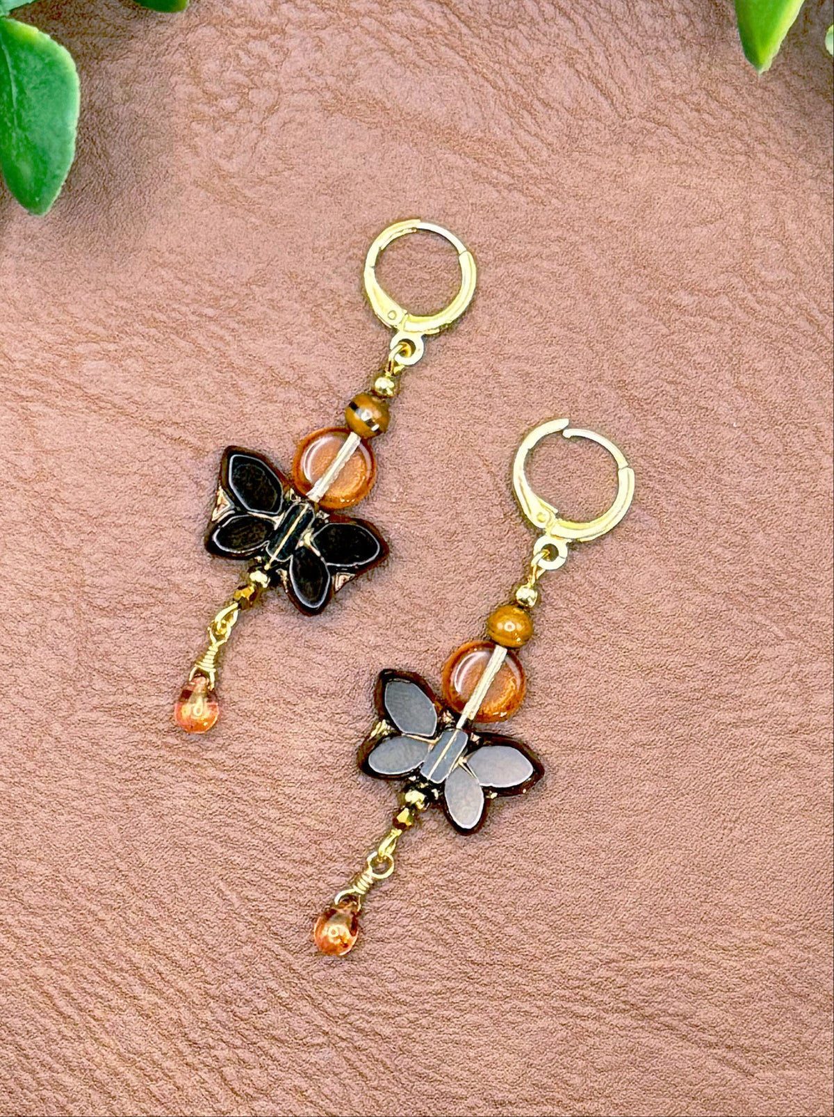 Handmade earrings with black butterfly charms, amber beads, and small dangling amber beads, displayed on a textured brown leather background with green leaves.