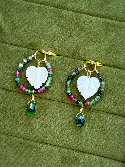 Handmade hoop earrings with gold wire, featuring a central white carved leaf charm, surrounded by small multicolored beads (green, pink, purple, black), and a dangling faceted green gemstone, displayed on an olive green textured background.