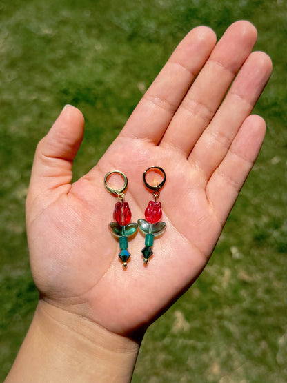 Handmade gold huggie earrings with red and green flower charms, against a blurred green grass background.