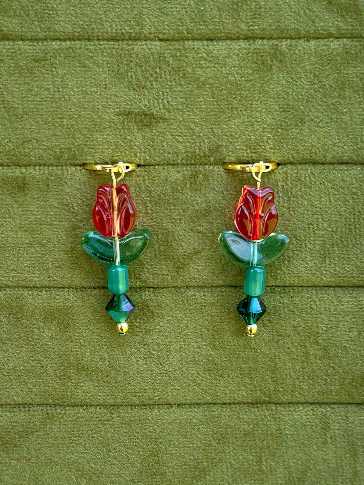 Handmade earrings with red flower-shaped beads, green leaves, and green faceted beads on gold huggies, displayed on a textured olive green background.