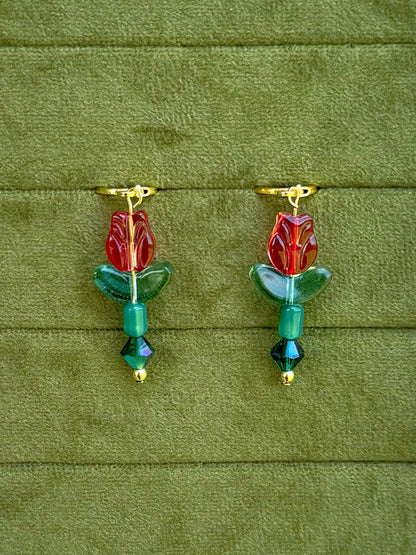 Handmade earrings with red flower-shaped beads, green leaves, and green faceted beads on gold huggies, displayed on a textured olive green background.