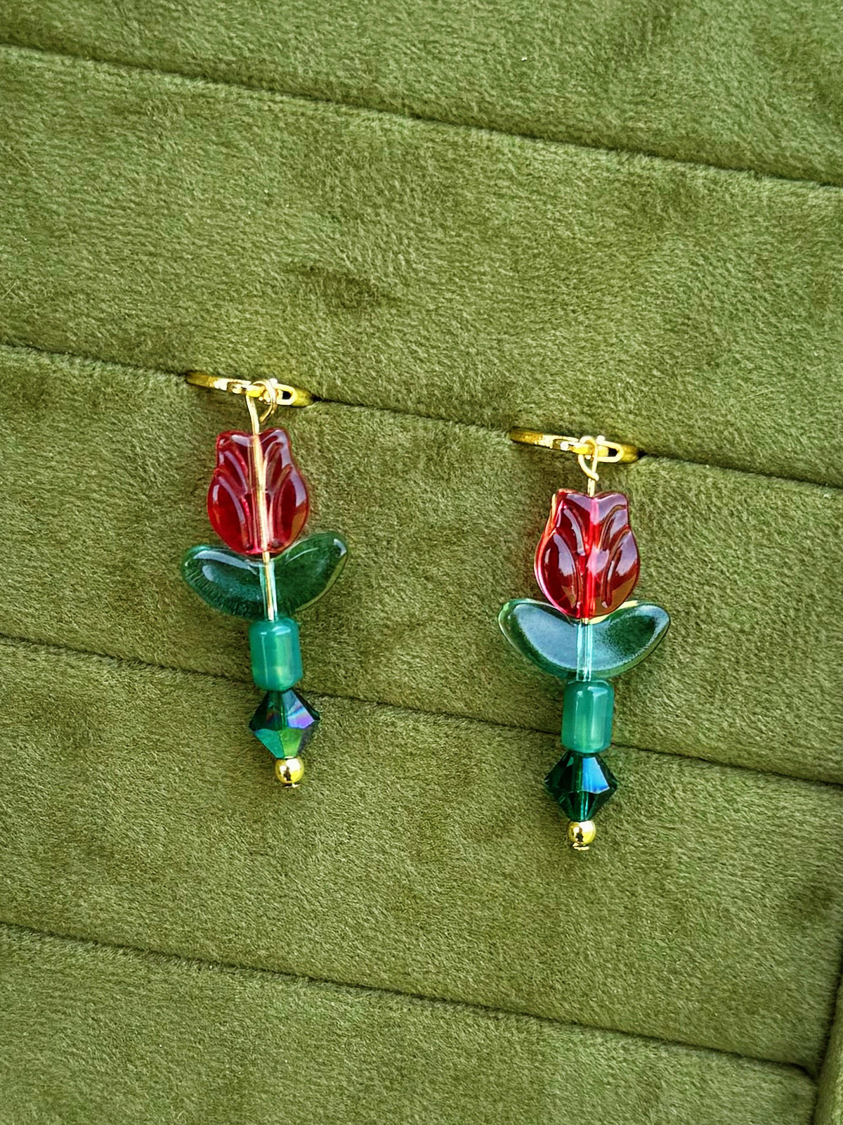 Handmade earrings with red flower-shaped beads, green leaves, and green faceted beads on gold huggies, displayed on a textured olive green background.