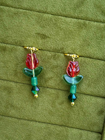 Handmade earrings with red flower-shaped beads, green leaves, and green faceted beads on gold huggies, displayed on a textured olive green background.