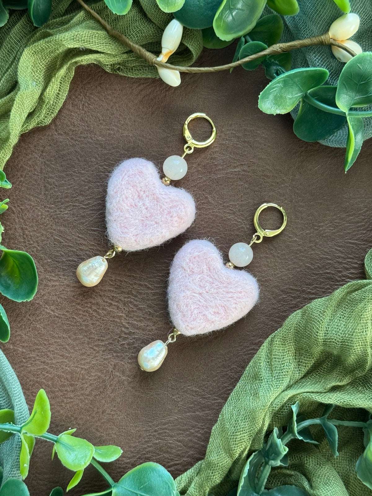 Handmade heart shaped needle felted earrings with gemstone beads on a natural background with green leaves around.