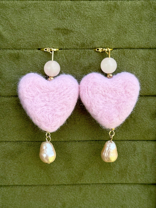 A pair of dangle earrings with fluffy light pink heart-shaped drops, a small white bead above, and a baroque pearl dangling below, shown on an olive green textured background.
