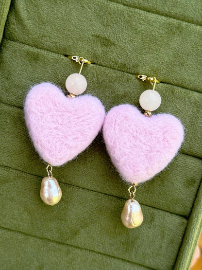 A pair of dangle earrings with fuzzy light pink heart-shaped drops, a small round white bead, and a dangling baroque pearl, displayed on an olive green velvet jewelry box.