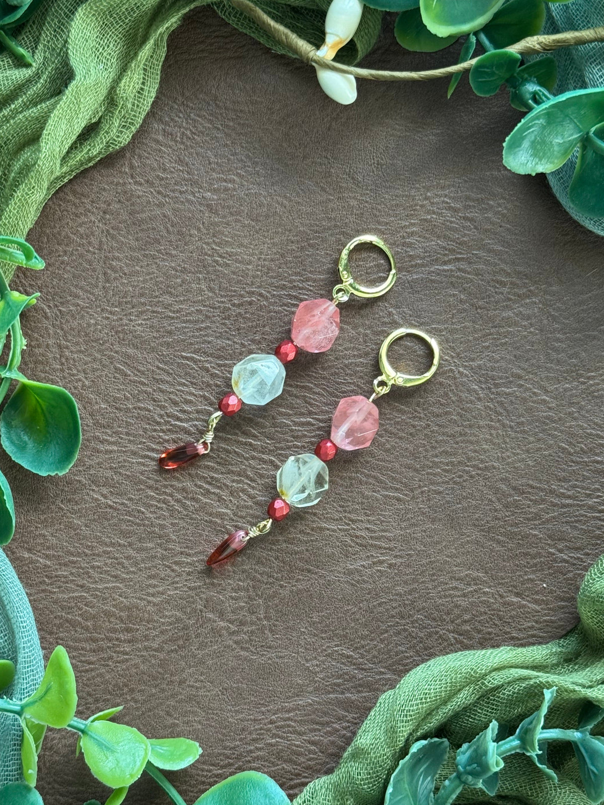 Handmade beaded earrings with gold huggies, pink and crystal beads, and red dangling beads, displayed on a natural surface surrounded by green leaves and fabric.