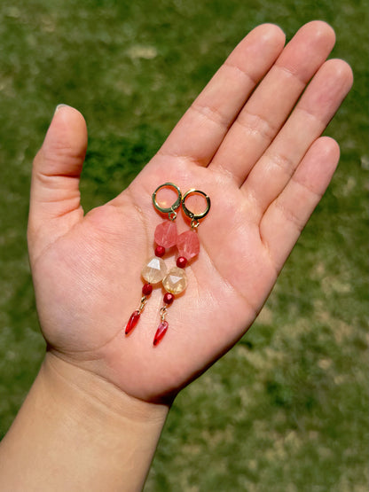 Handmade beaded earrings with gold huggies, pink and crystal beads, and red dangling beads, set against a blurred green grass background.