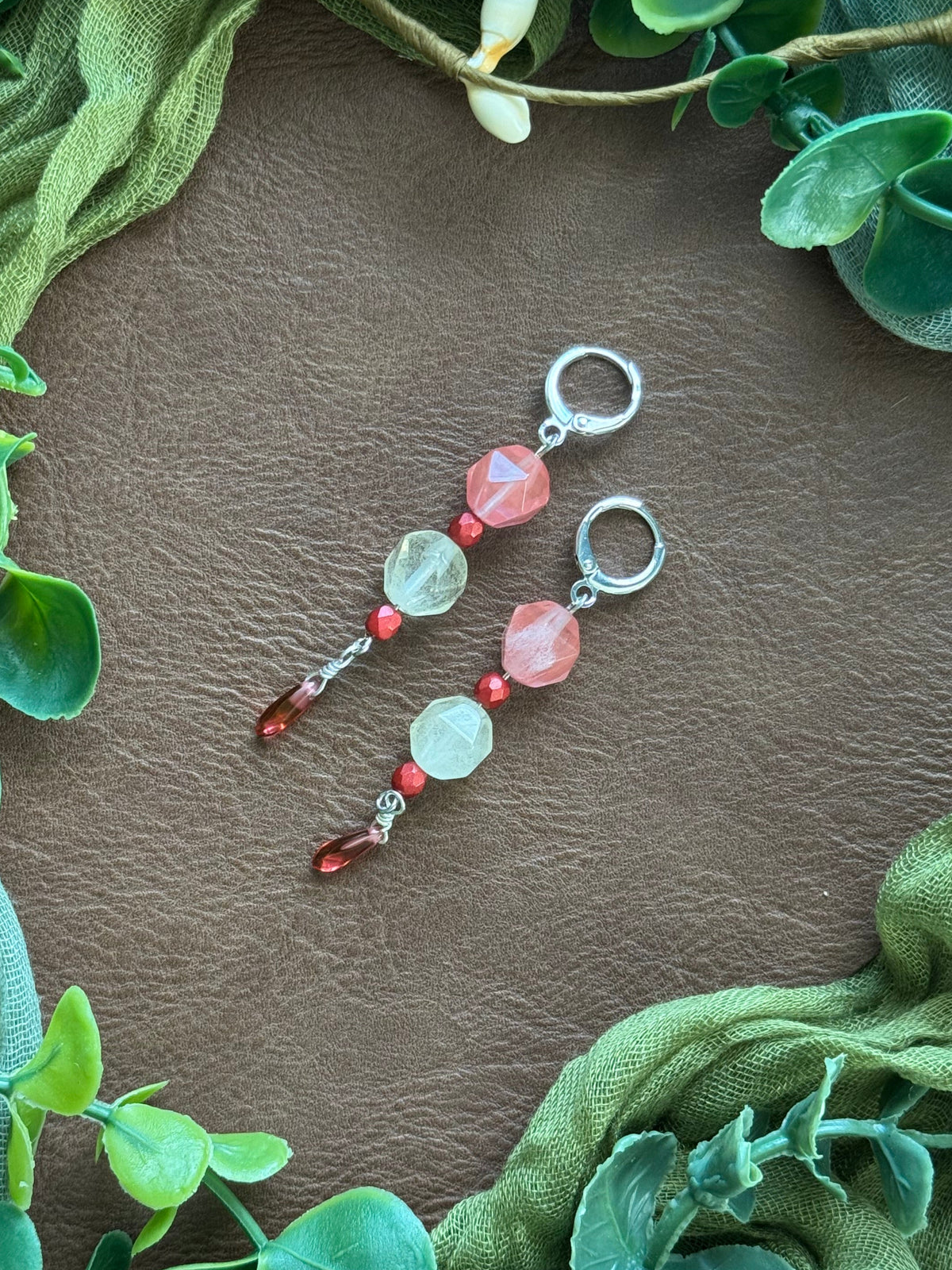 Handmade beaded earrings with silver huggies, pink and crystal beads, and red dangling beads, displayed on a natural surface surrounded by green leaves and fabric.