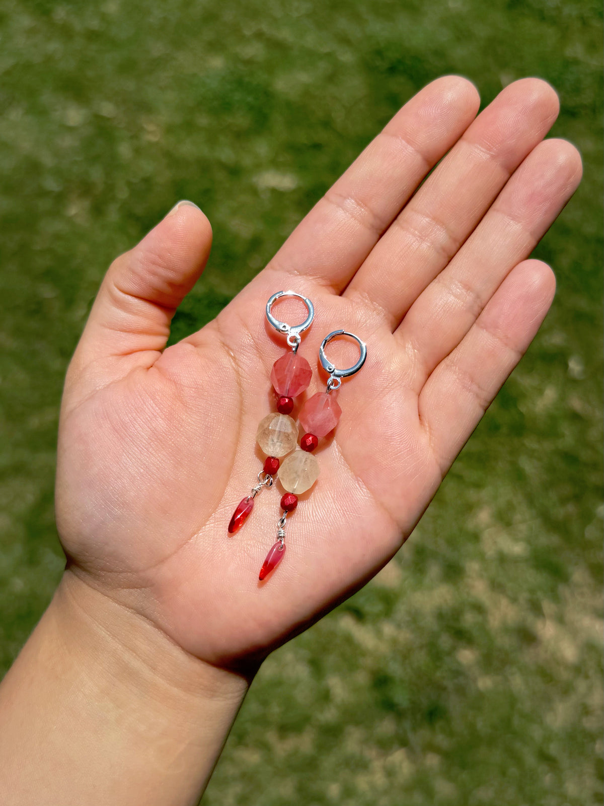 Handmade beaded earrings with silver huggies, pink and crystal beads, and red dangling beads, set against a blurred green grass background.