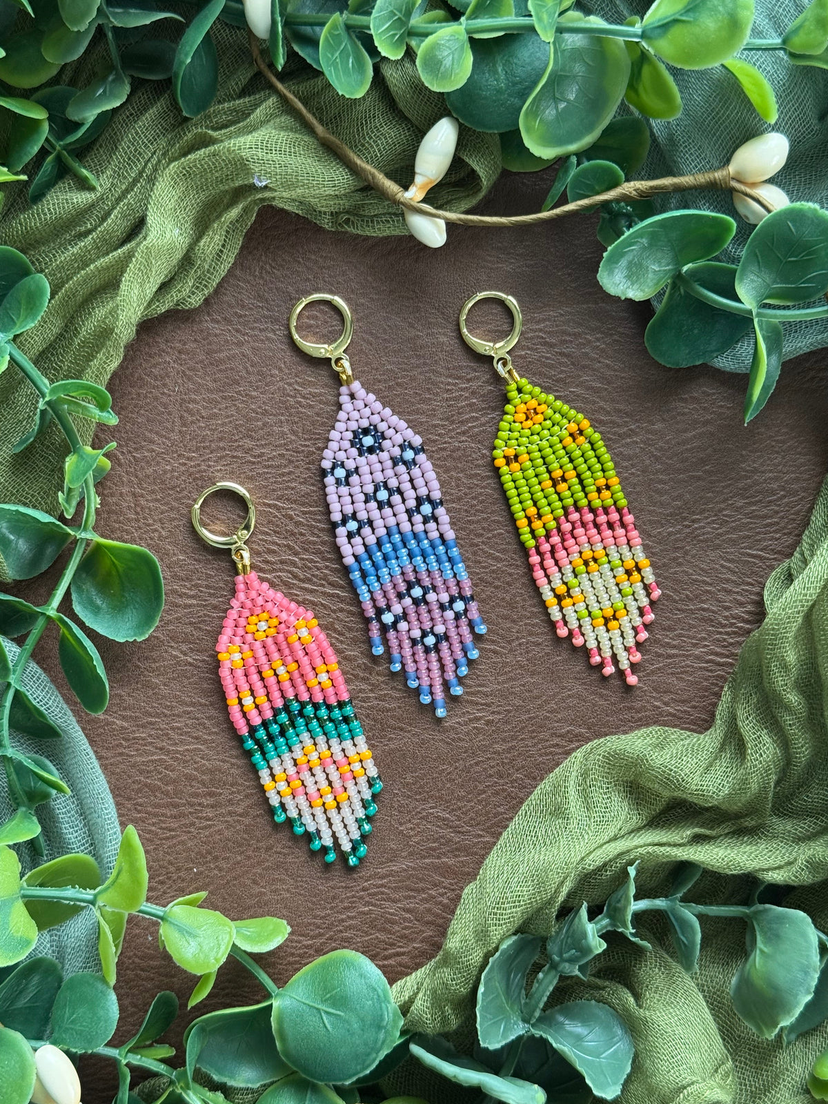Collection of handmade colorful beaded floral earrings using 11/0 glass seed beads on a natural surface surrounded by green leaves.