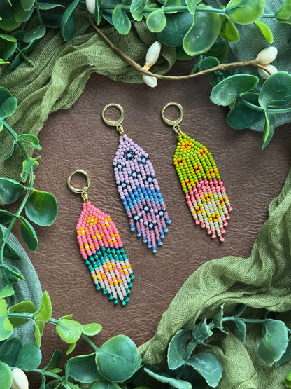 Collection of handmade colorful beaded floral earrings using 11/0 glass seed beads on a natural surface surrounded by green leaves.