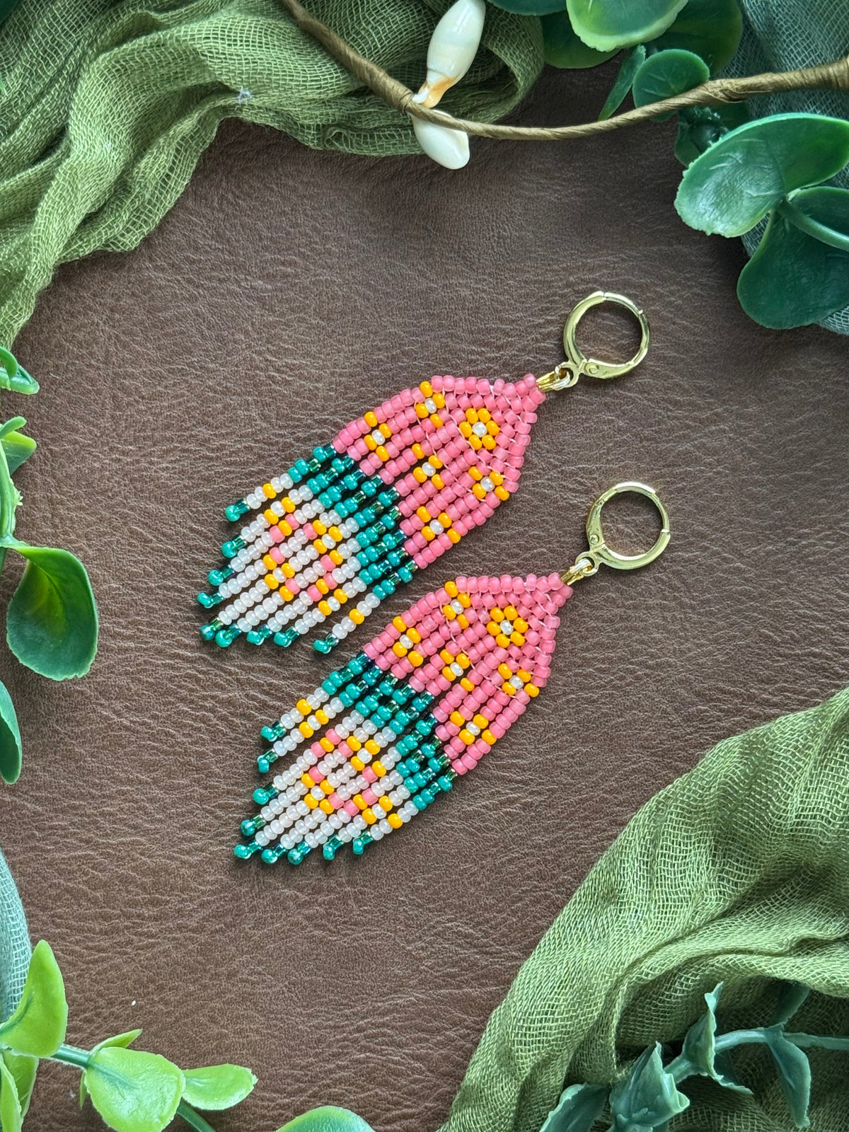 Handmade beaded earrings with a feather-like design in pink, teal, yellow, and white, hanging from gold huggies. They are displayed on a natural surface surrounded by green fabric and eucalyptus leaves.