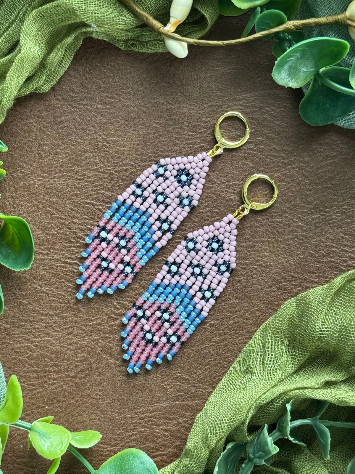 Handmade beaded earrings with a feather-like design in blue, purple, teal, and white, hanging from gold huggies. They are displayed on a natural surface surrounded by green fabric and eucalyptus leaves.