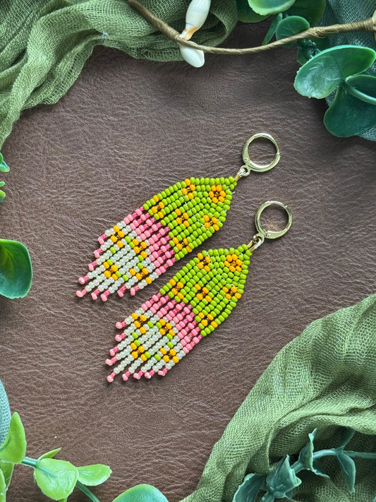 Handmade beaded earrings with green, pink, and yellow beads and gold huggies. Arranged on a natural surface surrounded by green leaves.