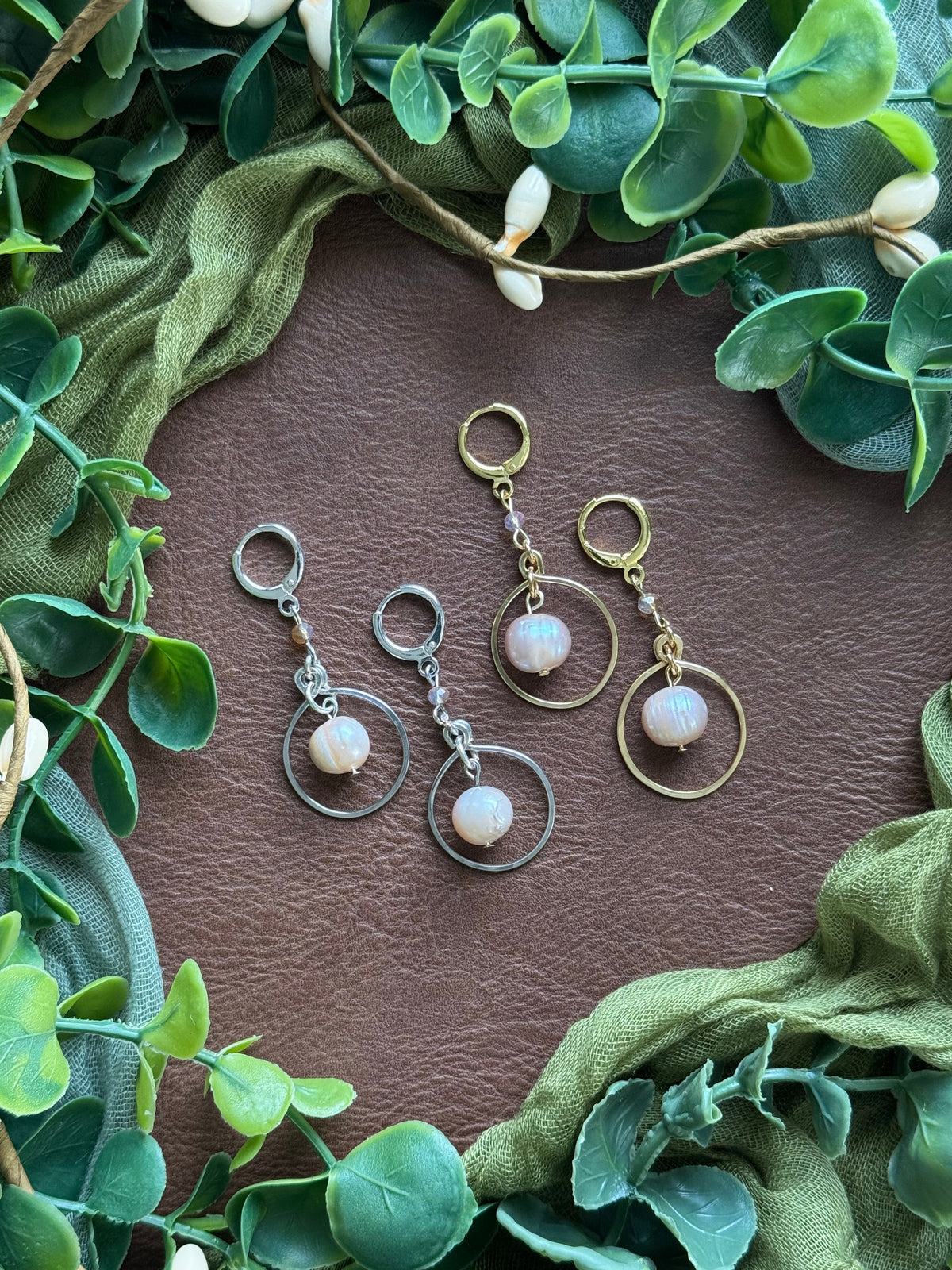 Collection of handmade beaded and wire-smithed earrings with pearl beads on a natural background with green leaves around.