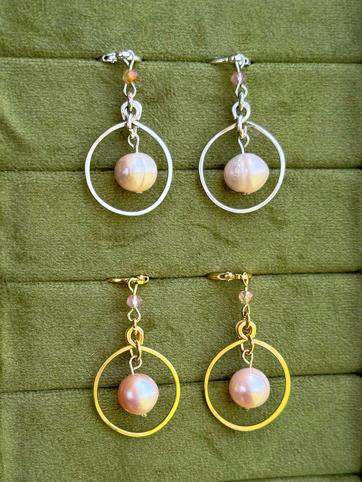 Handmade earrings displayed against a textured olive-green fabric background. The top pair features silver hoops with pale pink pearl beads, and the bottom pair features gold hoops with matching pale pink pearl beads.