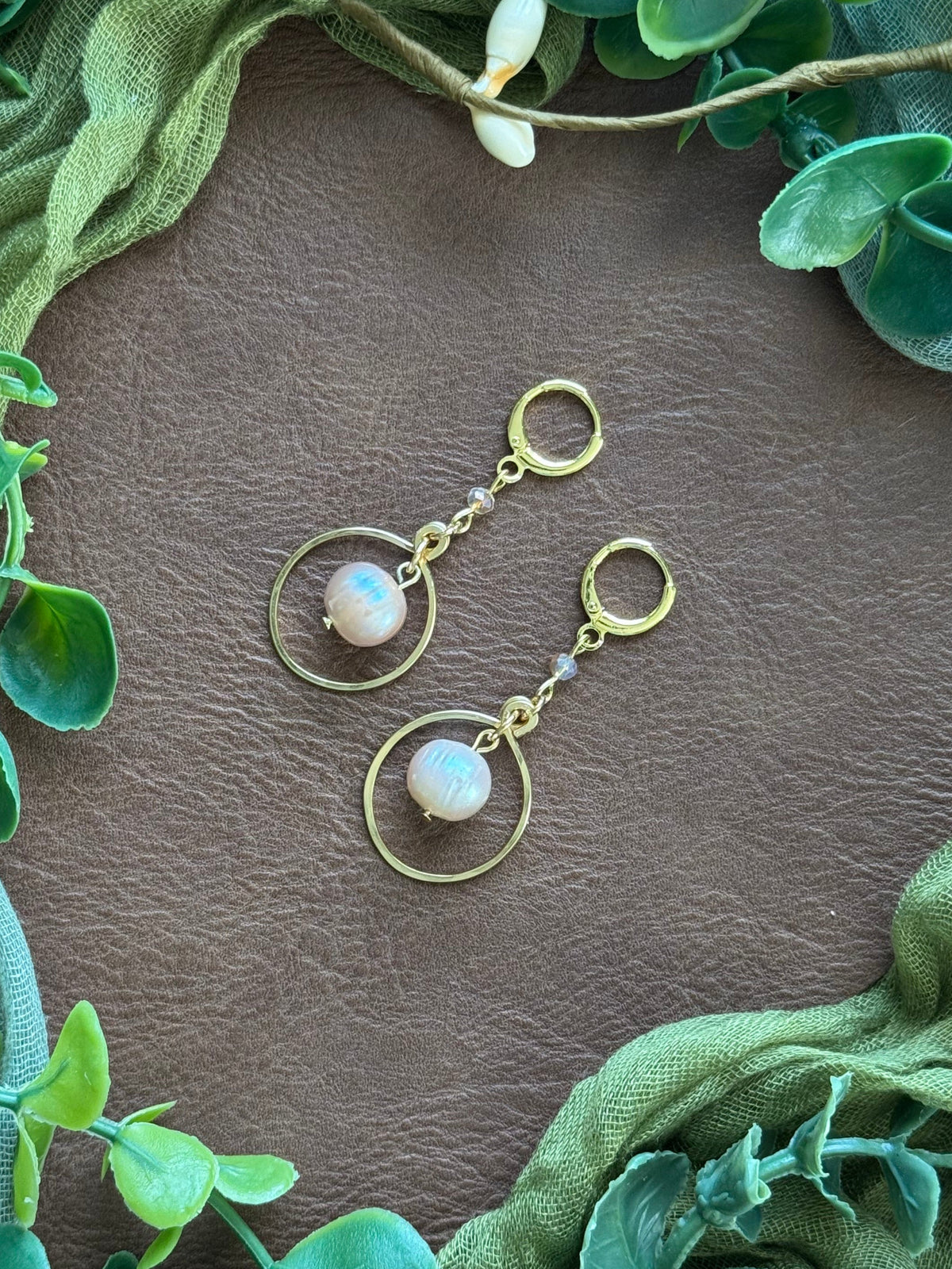 Handmade earrings with circular gold wire frames and freshwater pearl beads, arranged on a natural surface surrounded by green leaves.