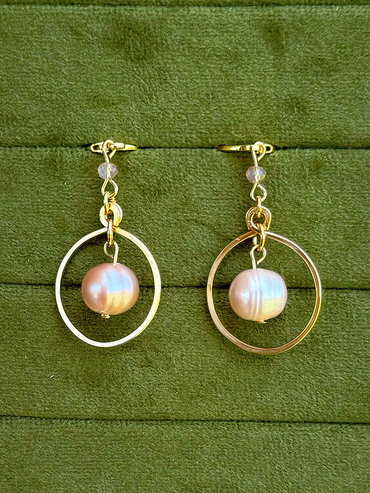 Handmade earrings with a single iridescent pinkish pearl hanging in the center of each hoop, displayed on a textured olive green background.