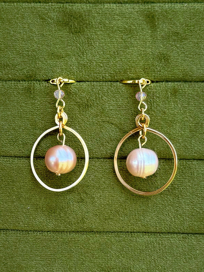 Handmade earrings with a single iridescent pinkish pearl hanging in the center of each hoop, displayed on a textured olive green background.