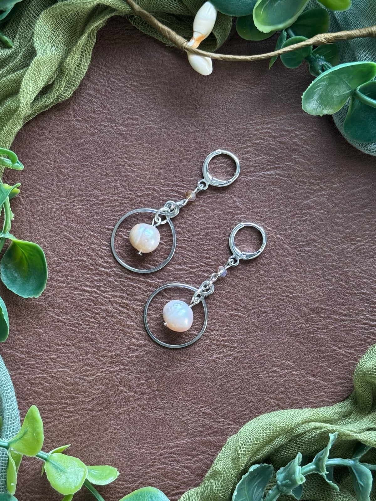 Handmade earrings with circular silver wire frames and freshwater pearl beads, arranged on a natural surface surrounded by green leaves.