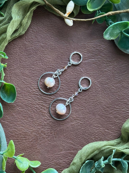 Handmade earrings with circular silver wire frames and freshwater pearl beads, arranged on a natural surface surrounded by green leaves.
