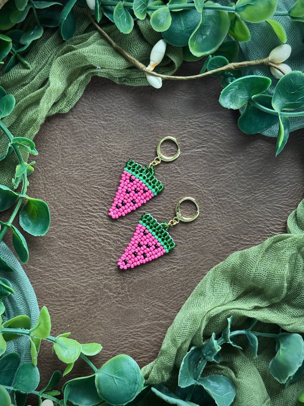 Handmade watermelon-themed beaded earrings using 11/0 glass seed beads on a natural surface with green leaves around.