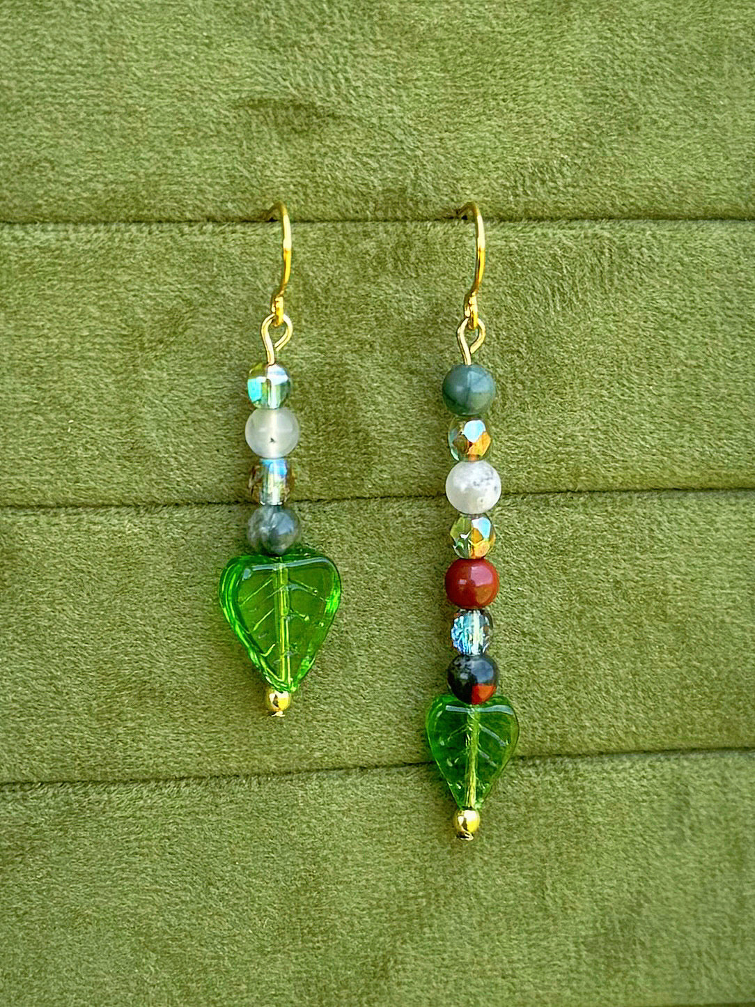 Handmade asymmetrical beaded earrings with gemstone and glass beads displayed on a textured olive green fabric background.