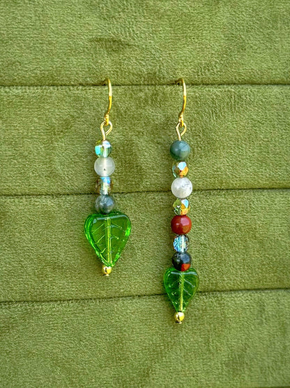 Handmade asymmetrical beaded earrings with gemstone and glass beads displayed on a textured olive green fabric background.