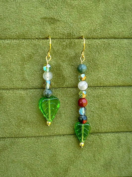 Handmade asymmetrical beaded earrings with gemstone and glass beads displayed on a textured olive green fabric background.