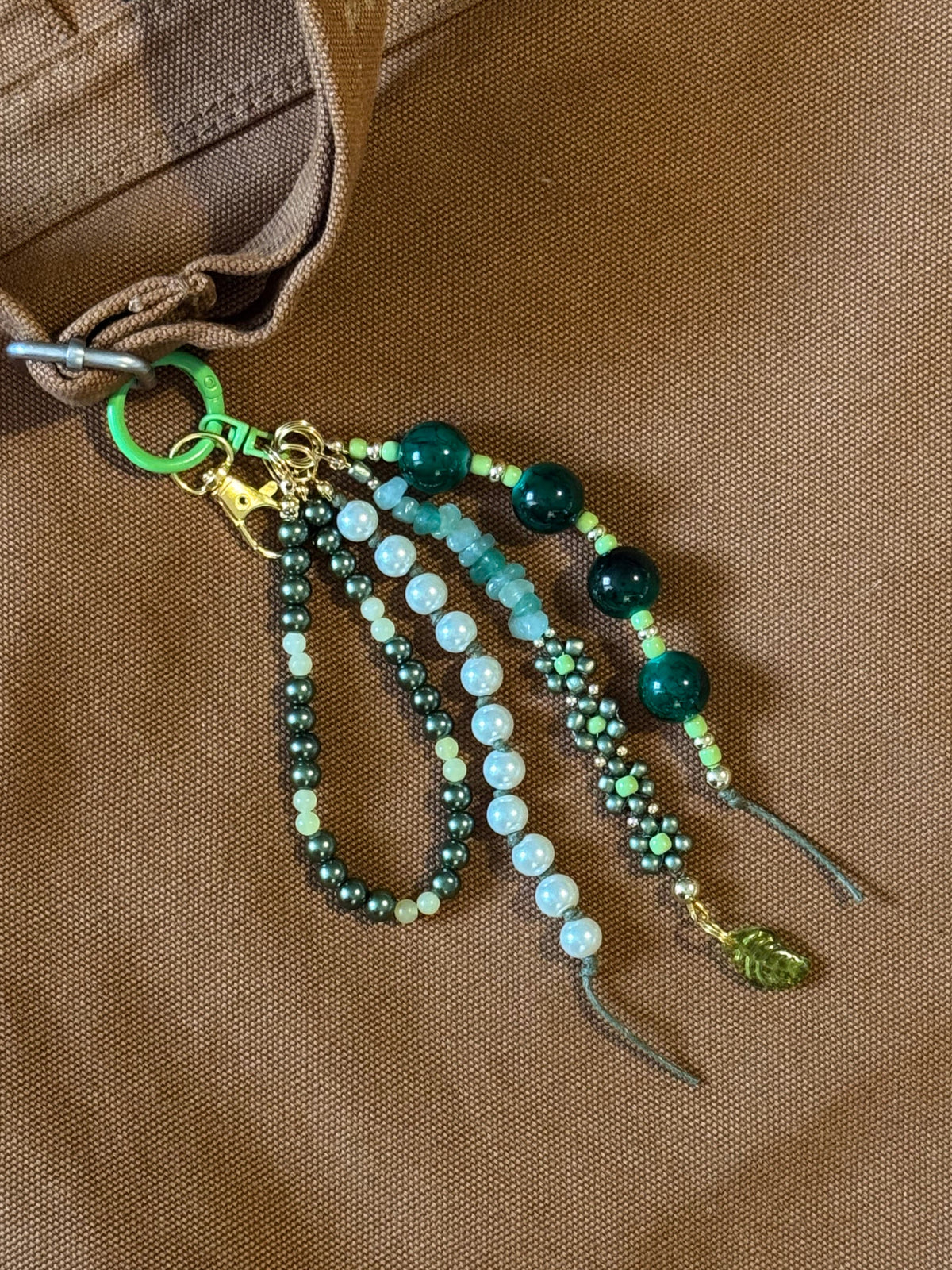 Handmade beaded keychain or bag charm with green and white beads, a gold clasp, and a green keyring, laid on a brown canvas surface.