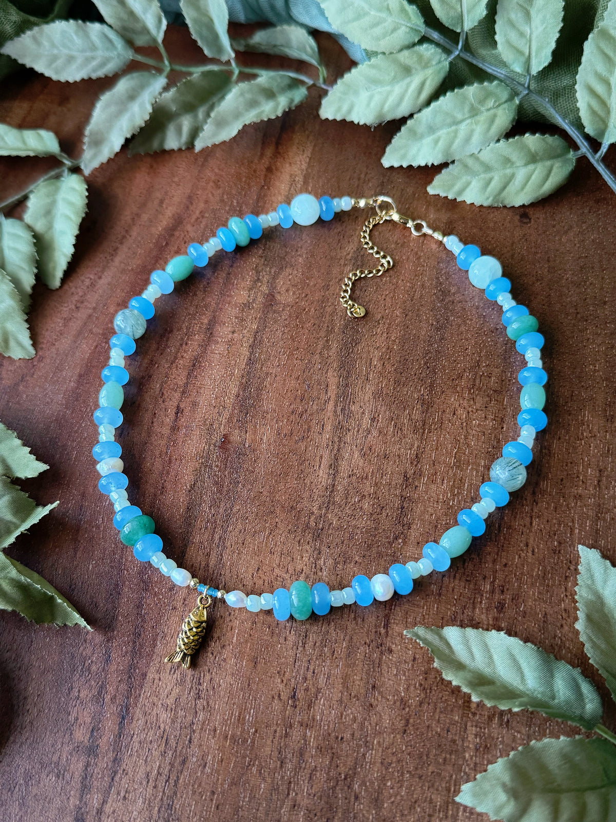 Necklace with blue beads and gold charm on a wooden surface with leaves