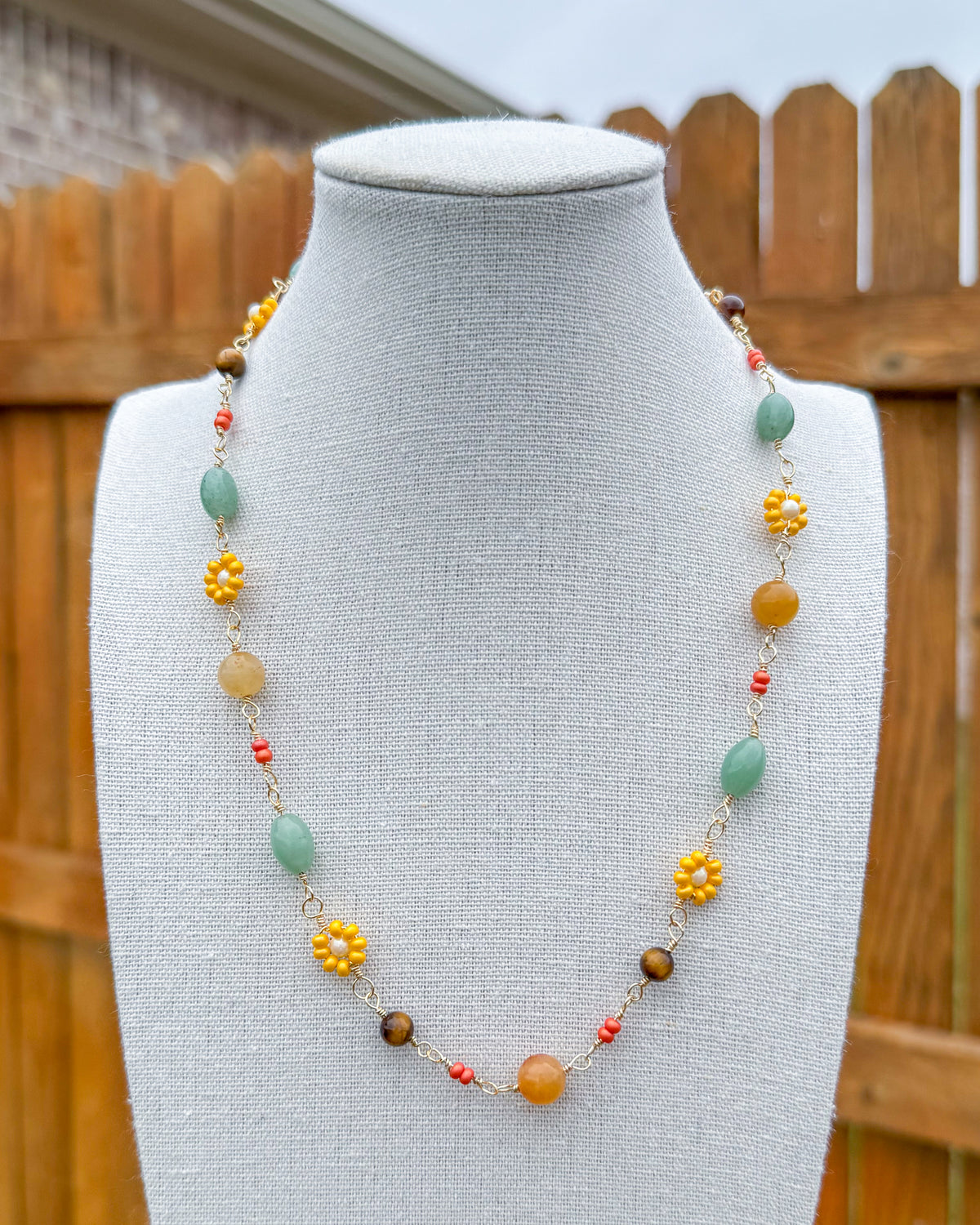 Handmade beaded necklace displayed on a mannequin bust, with a wooden fence and outdoor background. The necklace features a mix of green aventurine, yellow, orange, tiger's eye, and orange beads.