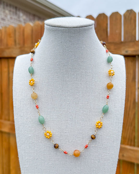Handmade beaded necklace displayed on a mannequin bust, with a wooden fence and outdoor background. The necklace features a mix of green aventurine, yellow, orange, tiger's eye, and orange beads.
