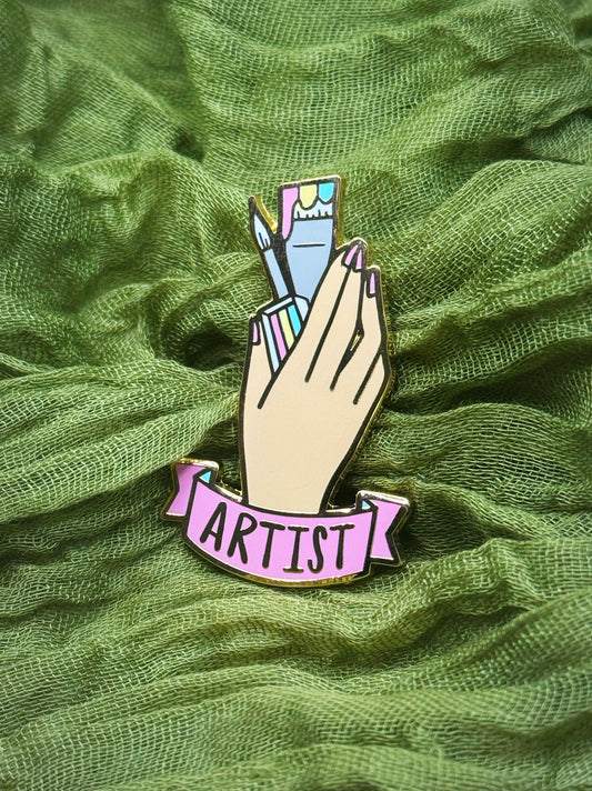Enamel pin with a hand holding colorful pencils and 'Artist' banner on green fabric