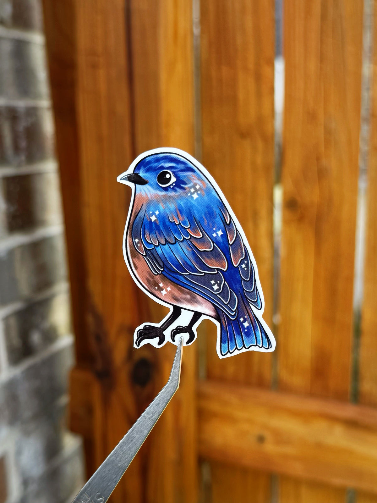 Die-cut sticker of a blue bird with brown and white speckled details, shown perched on a metal stand against a blurred wooden fence background.