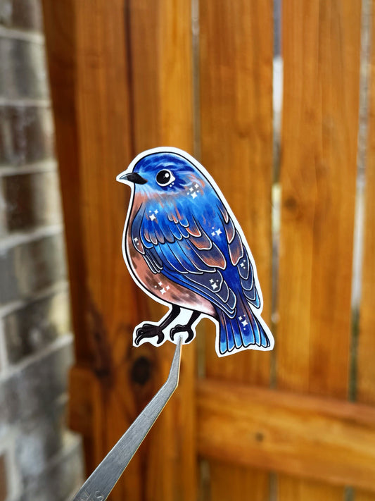 Die-cut sticker of a blue bird with brown and white speckled details, shown perched on a metal stand against a blurred wooden fence background.