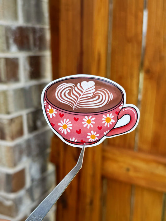 Die-cut sticker of a pink coffee cup with latte art and daisy patterns, held by tweezers against a blurred background of a wooden door and brick wall.