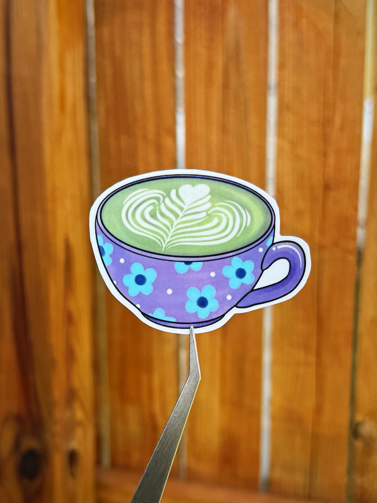 Die-cut sticker of a purple cup filled with matcha tea with latte art, held by metal tweezers against a blurred wooden background.