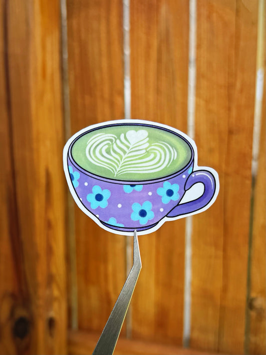 Die-cut sticker of a purple cup filled with matcha tea with latte art, held by metal tweezers against a blurred wooden background.